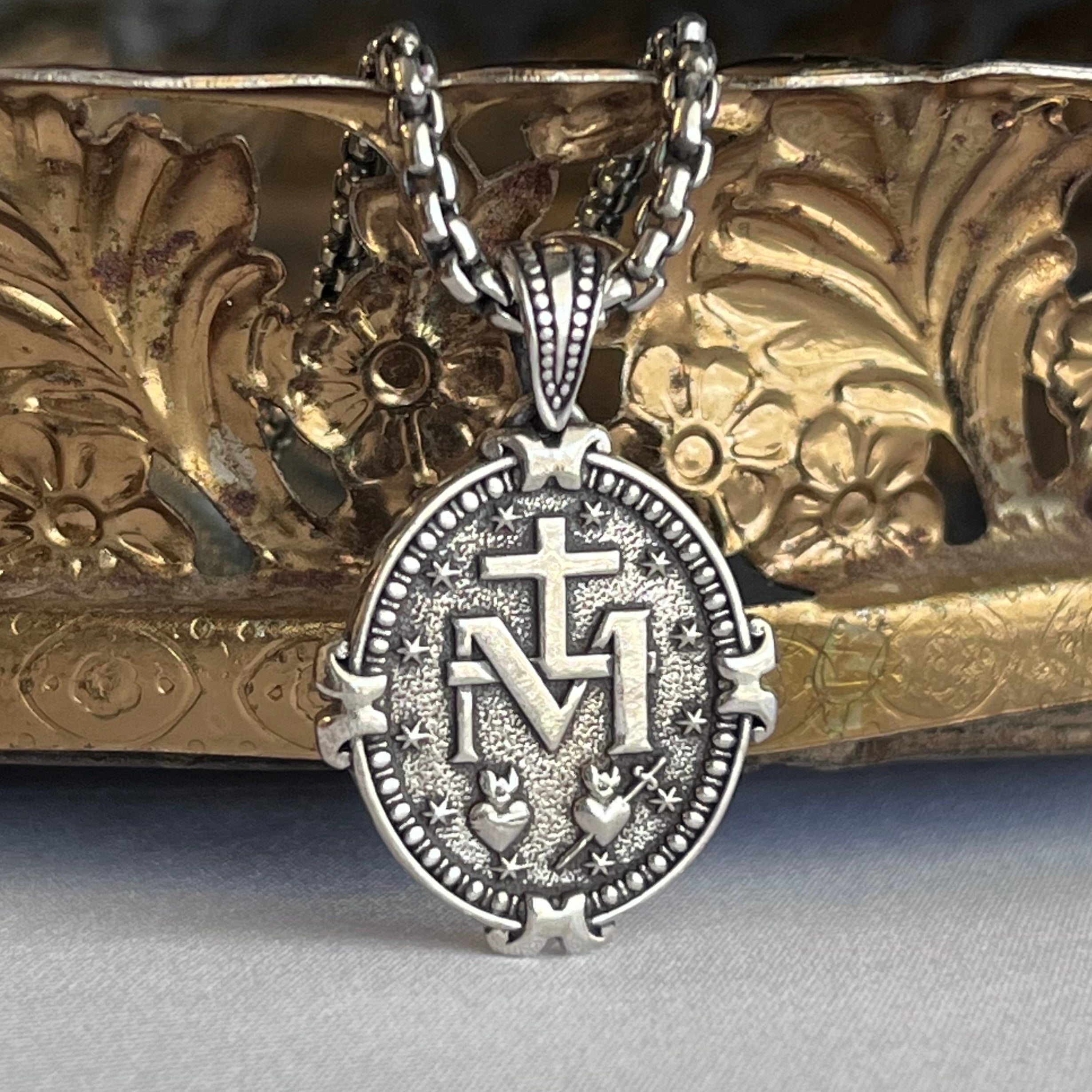 An oxidized sterling silver Miraculous Medal with a chain, displayed in front of a decorative gold-colored background.