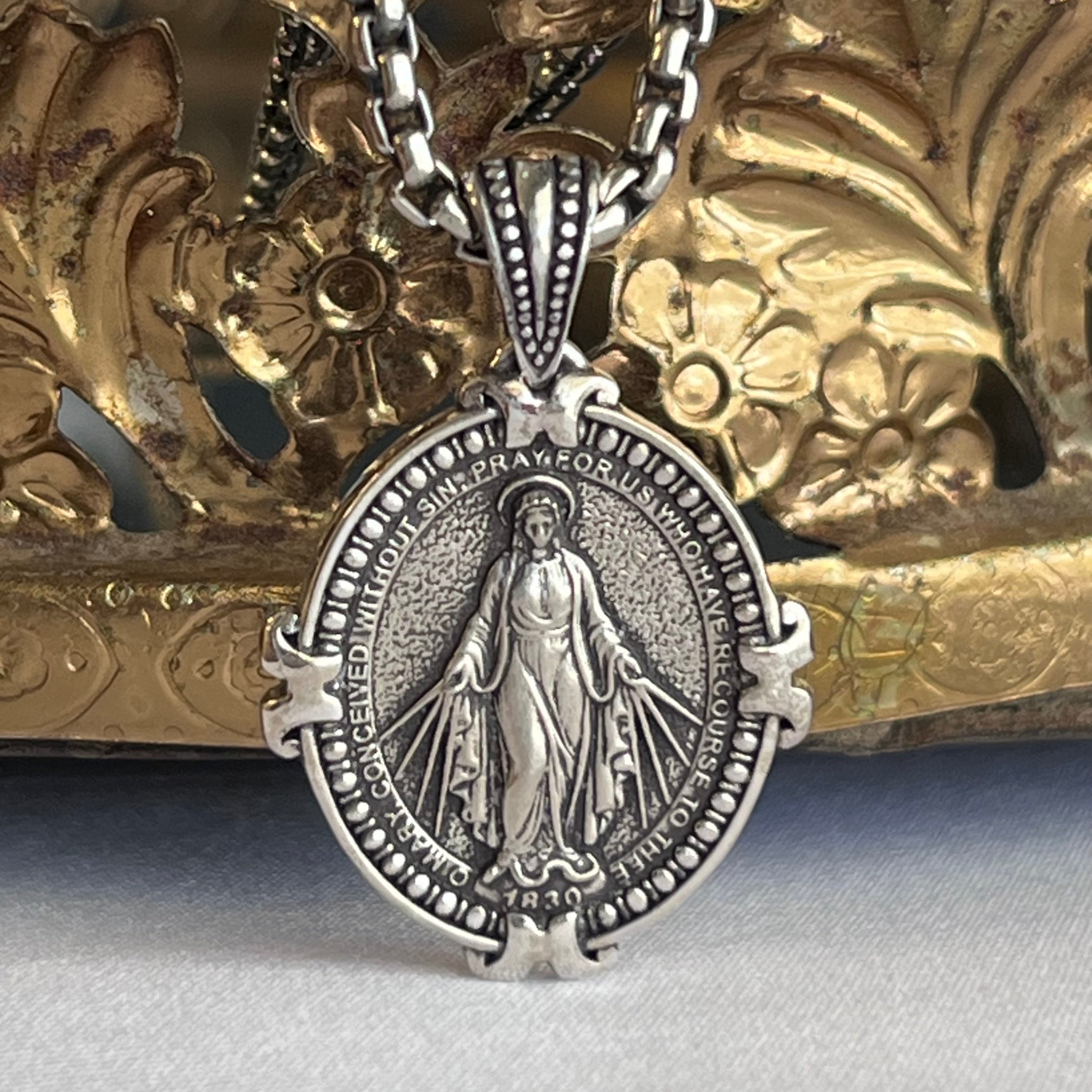 An oxidized sterling silver Miraculous Medal necklace featuring an image of the Virgin Mary standing with her hands extended. The necklace is displayed against an ornate golden background.