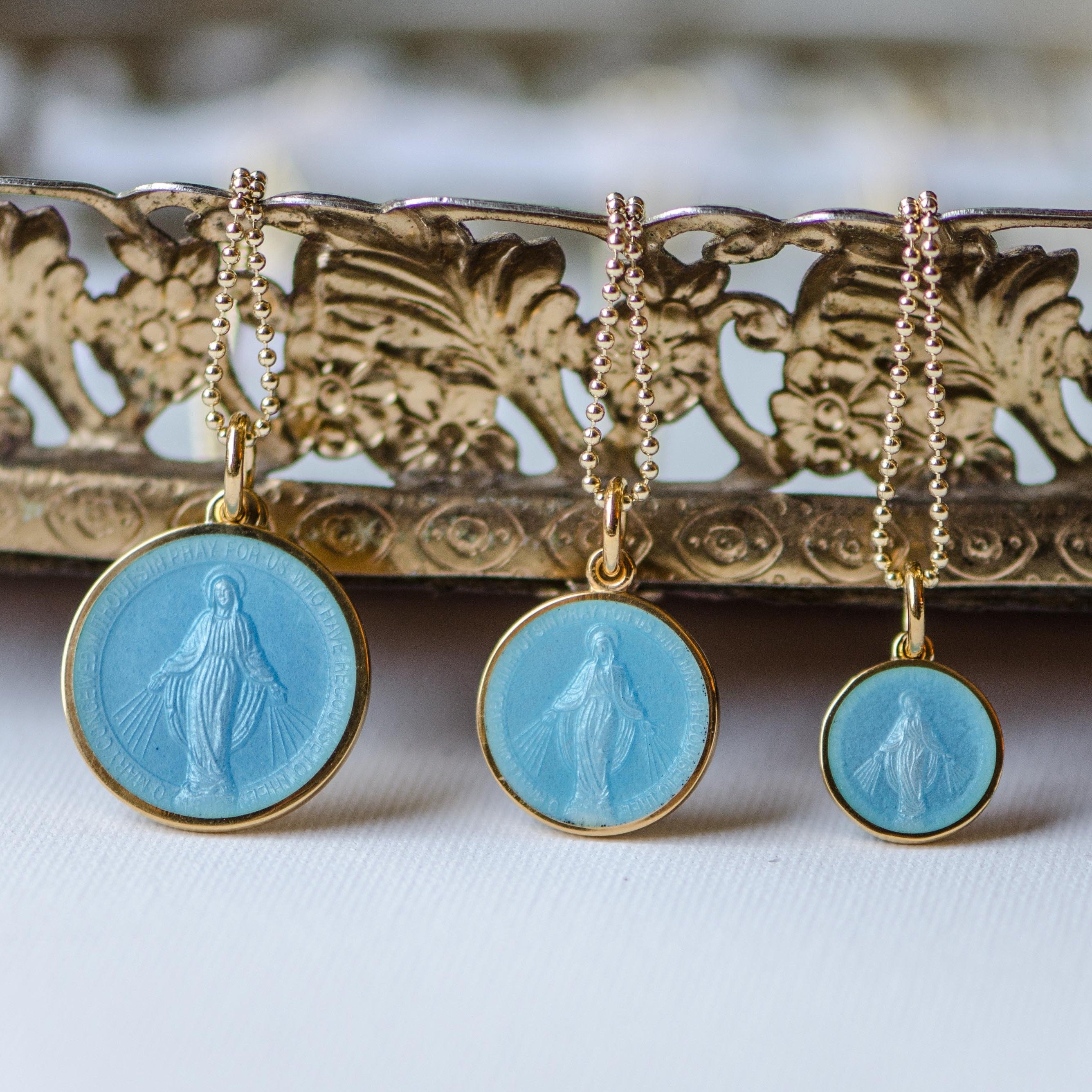 Three sizes of light blue enamel miraculous medal showing our lady. The medals are hanging from gold bead chains and posed in front of a decorative piece.