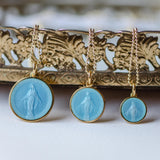 Three sizes of light blue enamel miraculous medal showing our lady. The medals are hanging from gold bead chains and posed in front of a decorative piece.