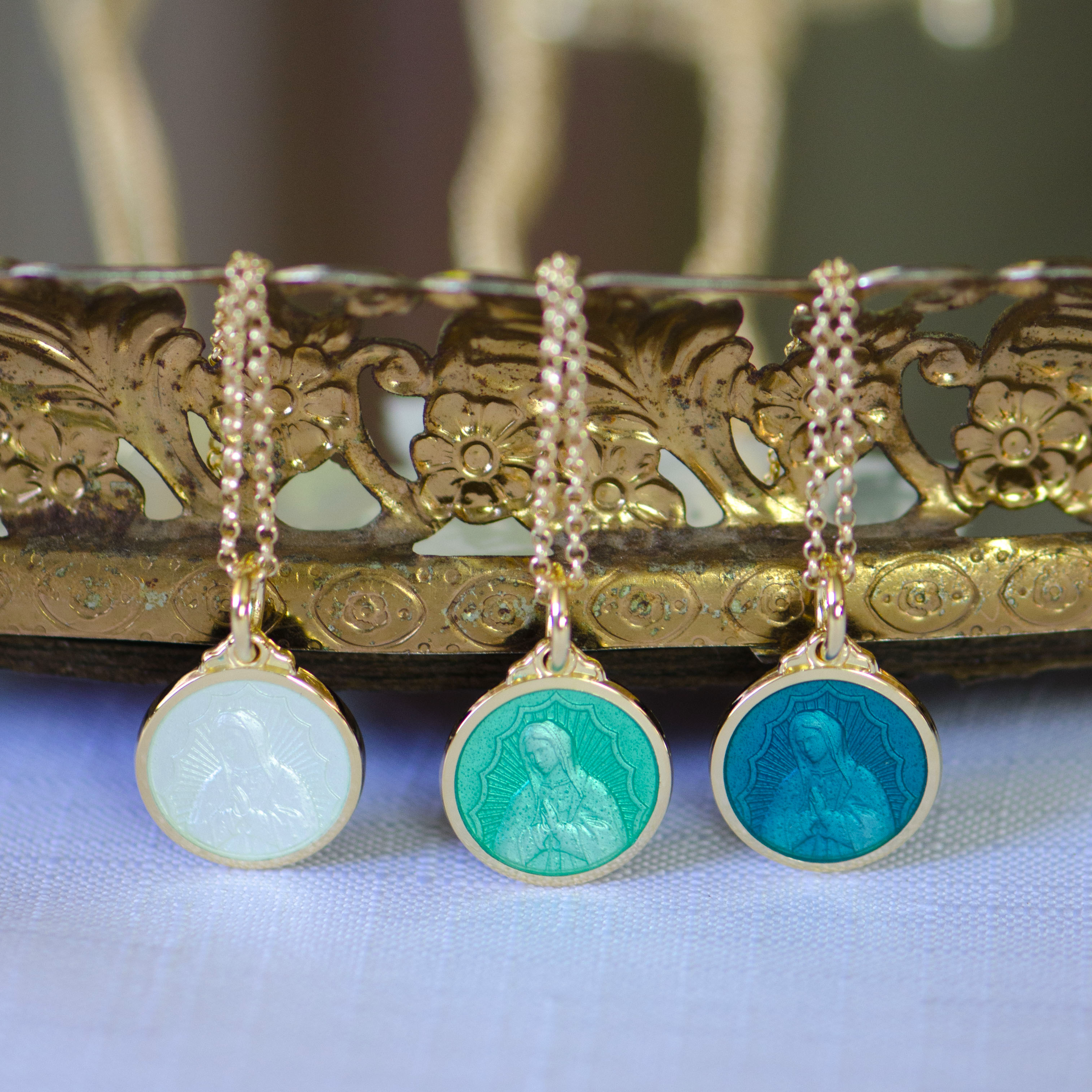 Three colored necklace pendants featuring Our Lady of Guadalupe highlighted with colored enamel overlay in white, teal, and light green