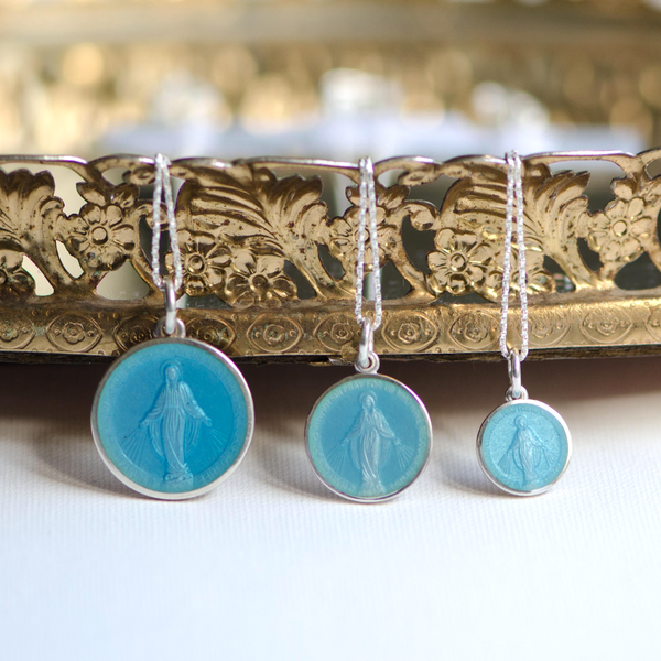 3 Miraculous medals of different sizes showing Our Lady behind light blue enamel.  The round medal is bordered by sterling silver and hung on a silver chain.
