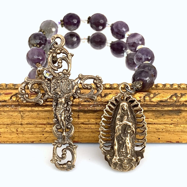 Rosary with Our Lady of Guadalupe bronze medal on right and open scroll crucifix on left with amethyst rosary beads in the background.