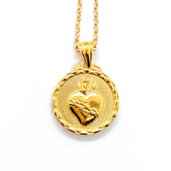 small gold necklace pendant hanging from a gold chain with the sacred heart of Jesus surrounded by a cut border around the outside of the pendant.